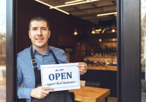 Small business owner smiling while holding open sign of restaurant reopening after lockdown quarantine due to coronavirus pandemic - Entrepeneur opening cafeteria activity to support local businesses.